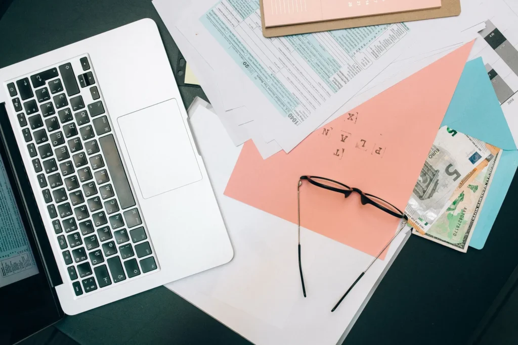 Laptop beside eyeglasses, paperwork and cash on a desk