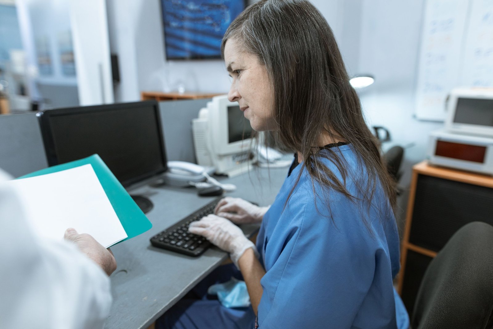 Healthcare staff member in scrubs typing at a hospital office workstation as a colleague holds a file
