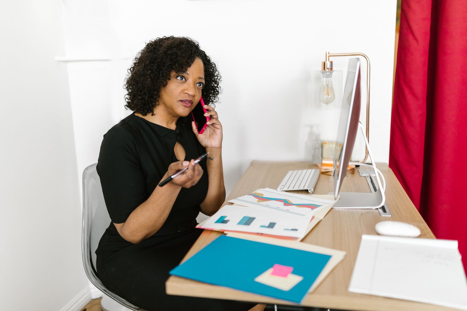 Office staff member on a phone call at her desk, pen in hand, with paperwork and a computer