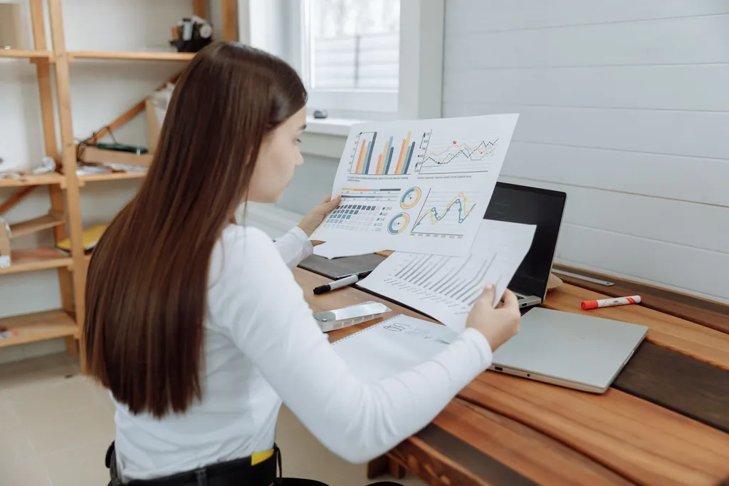 Woman at a desk reviewing printed charts and reports beside a laptop
