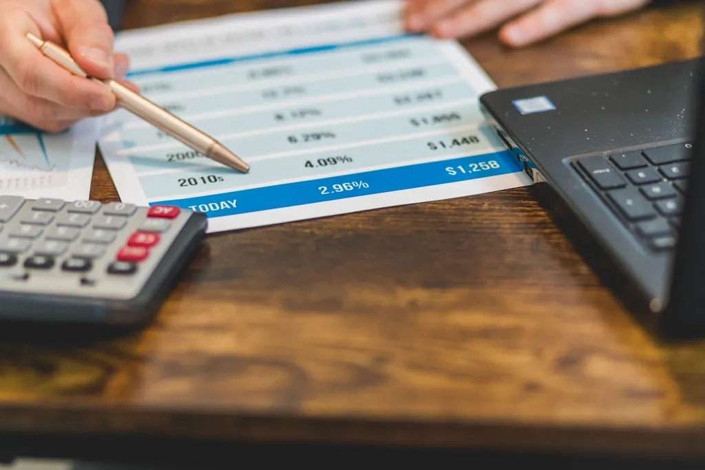 Hand pointing a pen at a printed rate table beside a calculator and laptop