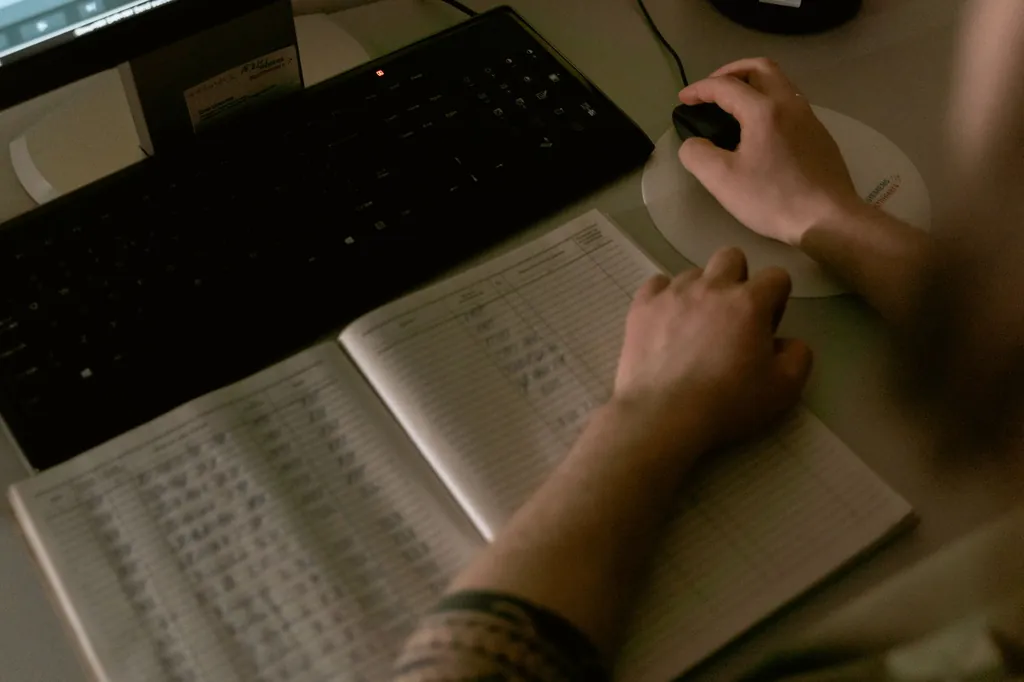 Hands at a keyboard and mouse beside an open notebook of handwritten entries