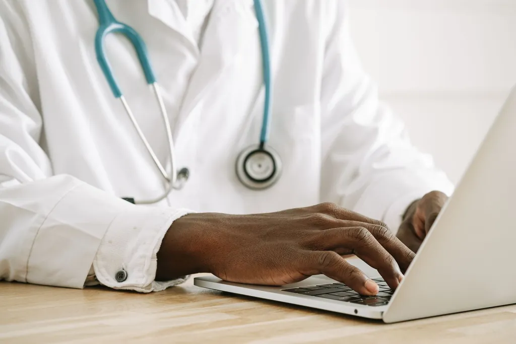 Clinician in a white coat with a stethoscope typing on a laptop