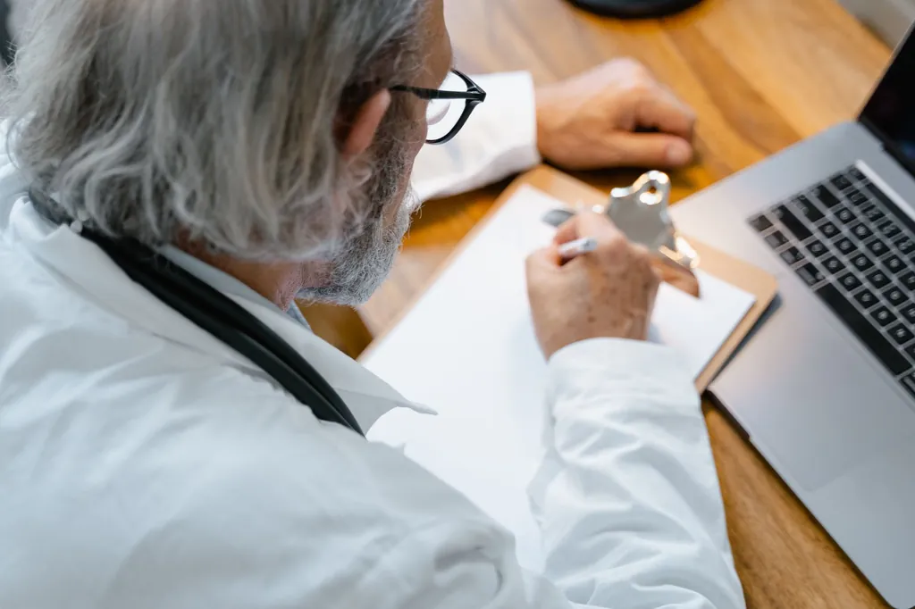 Physician writing notes on a clipboard at a desk beside a laptop