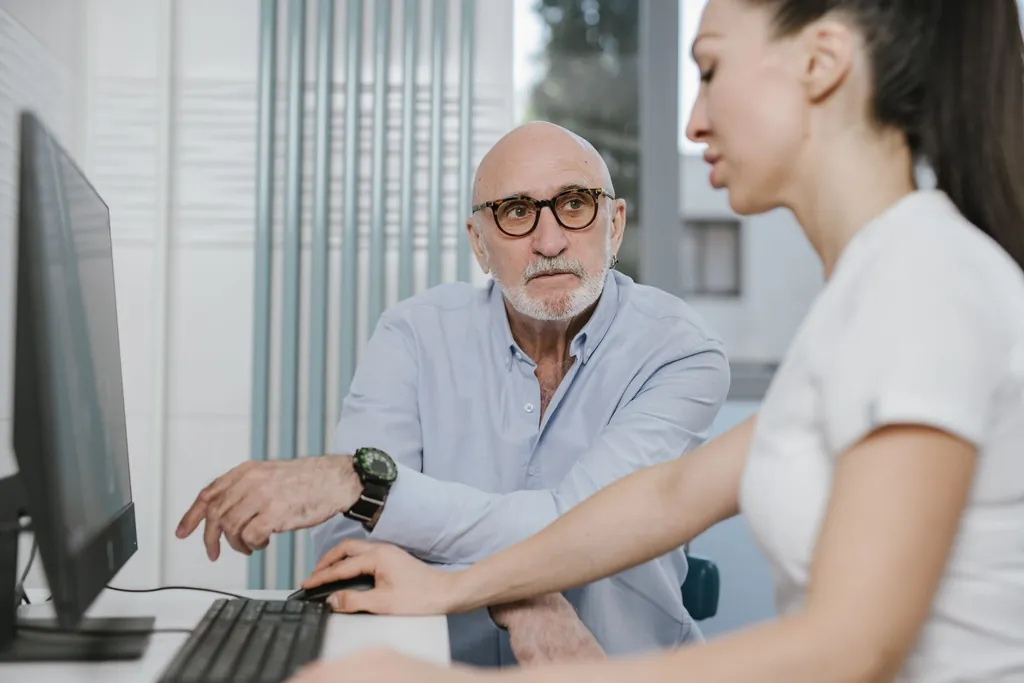 Older man in glasses talking with a woman at a computer desk
