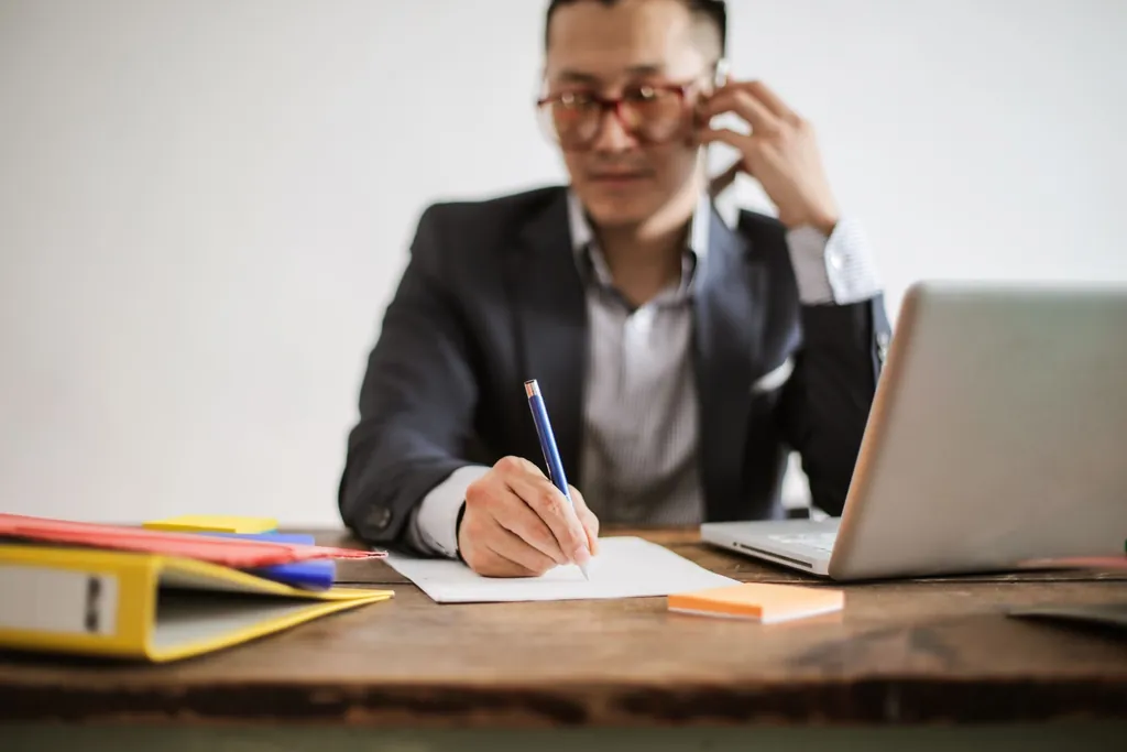 Man on a phone call taking notes at a table with his laptop