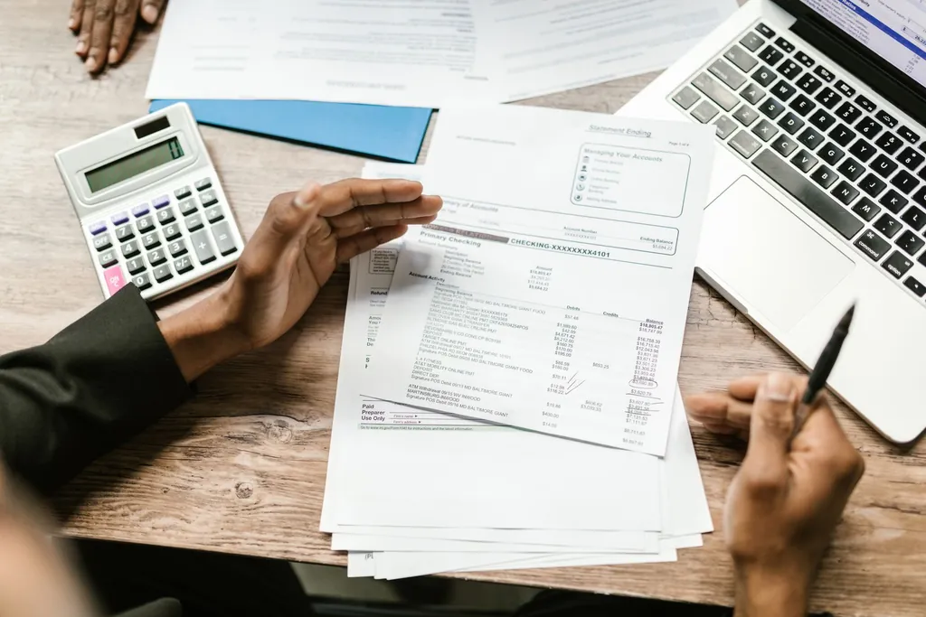 Hands going through payment statements beside a calculator and laptop